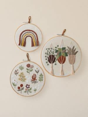 Three round embroidered wall hangings featuring a rainbow, hanging plants with tassels, and assorted florals in neutral tones on a light wall.