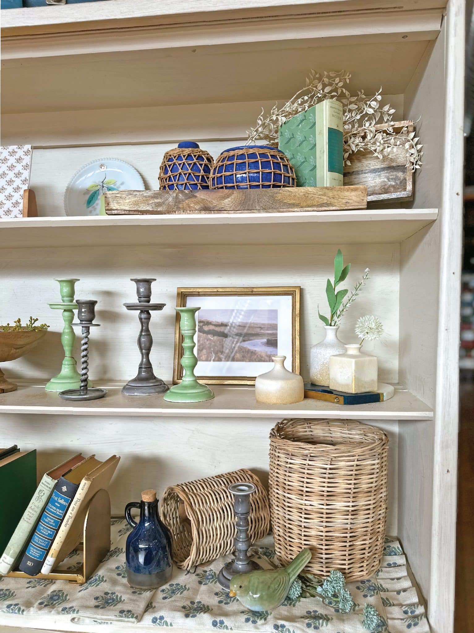 Front view of shelves styled with an eclectic country cottage feel, featuring blue and green tones, neutral textures, candlesticks, vases, baskets, landscape frames, and books for a timeless look.