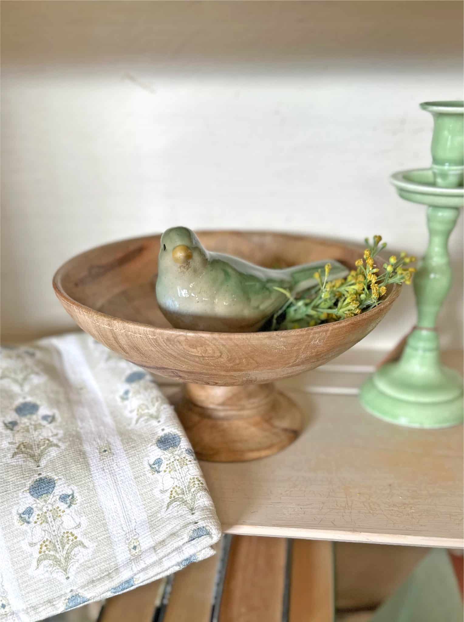 Cottagecore green bird figurine nesting inside of a neutral wood pedestal footed bowl. All resting in a a hutch on a neutral and blue cottage floral towel.